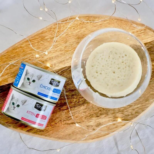 Kit Cat Kitten Chicken Mousse cans on a wooden platter with a bowl of mousse, showcasing grain-free cat food.