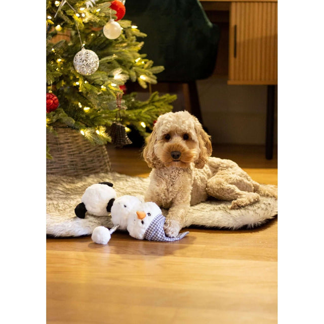 Dog playing with Snoop Snowman toy by Christmas tree, showcasing festive atmosphere and plush fun.
