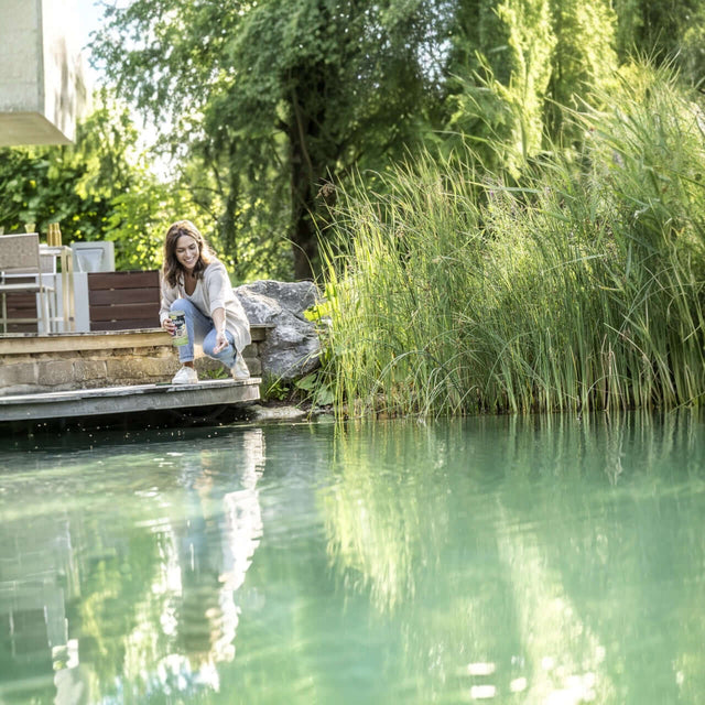 Woman feeding fish by a tranquil pond, surrounded by lush greenery and sunlight.