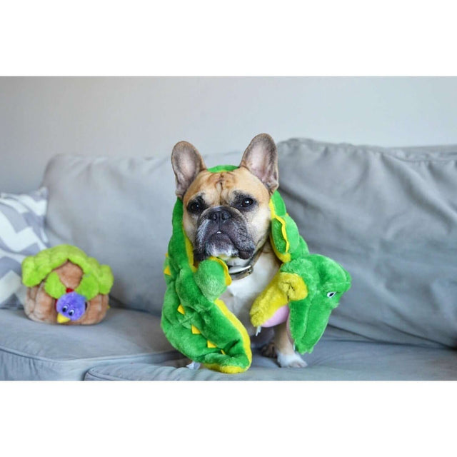 French Bulldog wrapped in a green plush toy blanket, sitting on a couch with squeaky toys nearby.