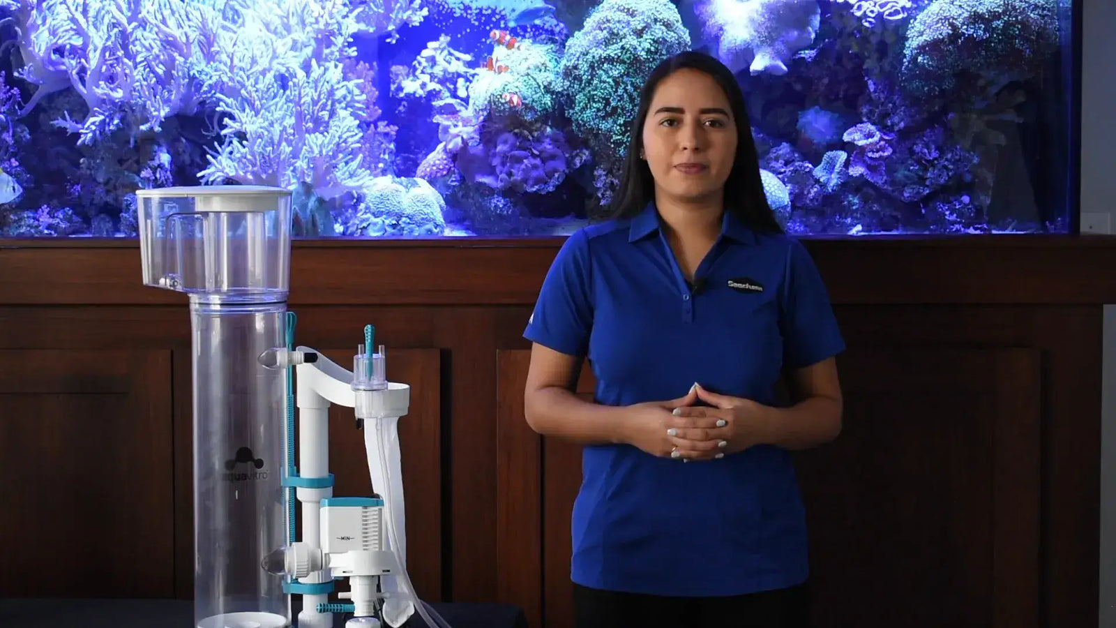 A woman standing next to the AquaVitro Division protein skimmer in front of a coral aquarium backdrop.