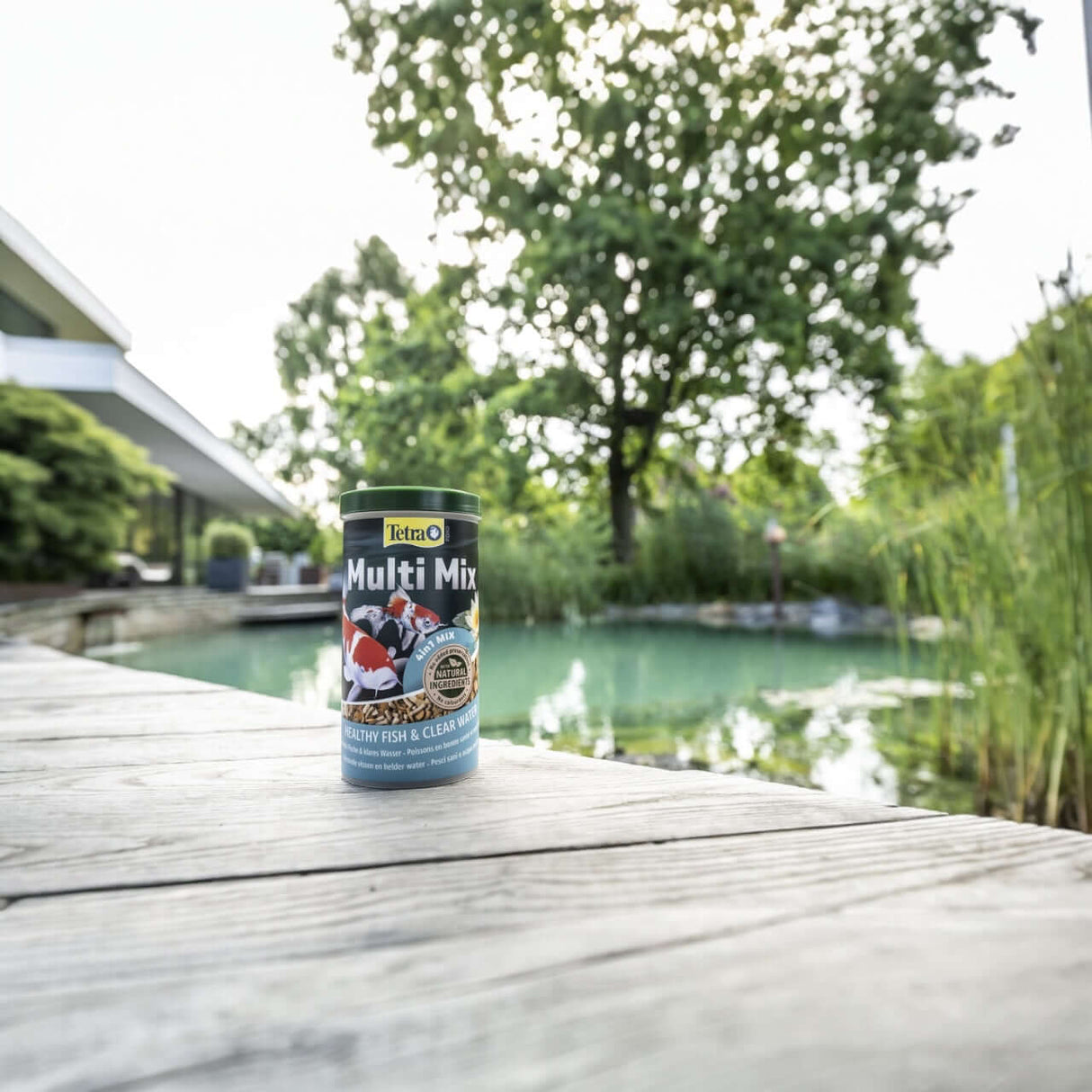 Tetra Pond Multi Mix 1L can on wooden deck by a serene pond, ideal food for all pond fish and enhances water quality.