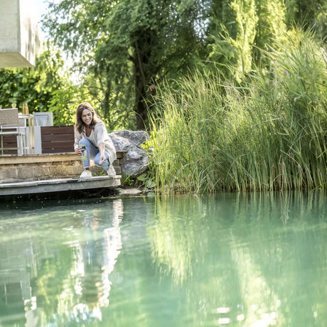 Woman feeding fish by a tranquil pond, surrounded by lush greenery and sunlight.